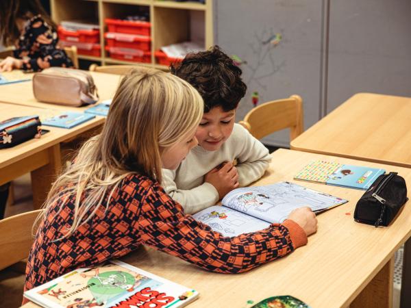 twee leerlingen zitten aan hun schoolbank en lezen samen in een boek